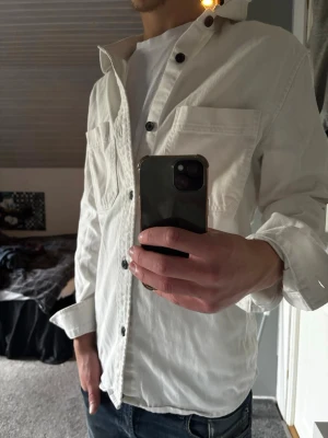 Vit Overshirt  - Stilren vit skjorta med svarta knappar och bröstficka. Skjortan har lång ärm och klassisk krage, perfekt att bära öppen över en t-shirt för en avslappnad look. Passar till jeans eller chinos för en enkel men snygg outfit.