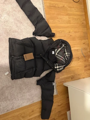 Svart pufferjacka från Burberry som bilar också väst  - Säljer en svart pufferjacka från Burberry med klassiskt rutigt foder i huvan. Jackan har dragkedja och knappar framtill, två snedställda fickor med dragkedja och en huva som ger extra värme. Perfekt för kalla dagar och riktigt snygg streetstil.