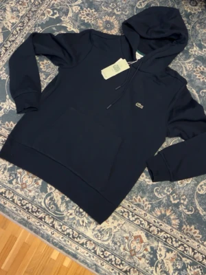 Mörkblå hoodie från Lacoste - Snygg mörkblå hoodie från Lacoste med klassisk passform. Hoodien har dragsko i huvan, stor magficka och den ikoniska krokodilloggan på bröstet. Tillverkad i mjuk bomull som känns skön mot huden. Perfekt för en chill och stilren look.