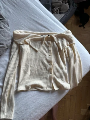 Off shoulder ribbad cardigan cremevit - Trendig off shoulder cardigan i ribbad cremevit trikå med stora kragar och knappar framtill. Långärmad och figurnära modell som ger en cool och stilren look. Perfekt att styla med jeans eller kjol för en snygg outfit.