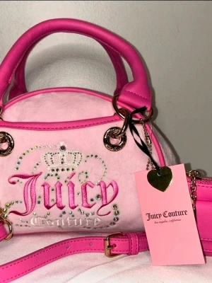 Juicy Couture rosa handväska - Säljer en ikonisk rosa handväska från Juicy Couture i mjuk velour med detaljer i skinnimitation. Väskan har broderad logga, glittrande stenar och guldfärgade metalldetaljer. Två handtag och avtagbar axelrem. Perfekt för dig som älskar Y2K-stil!