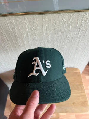 Grön Oakland Athletics keps New Era - Mörkgrön keps från New Era med Oakland Athletics 'A's' broderat i vitt framtill och MLB-logga på sidan. Klassisk basebollmodell med böjd skärm och broderade ventilationshål. Perfekt för dig som gillar sportig streetstyle. 60.6 cm i storlek