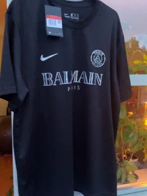 Svart PSG x Balmain t-shirt Nike - Snygg svart t-shirt från Nike i samarbete med PSG och Balmain. T-shirten har vita loggor och text framtill, rund hals och korta ärmar. Perfekt för dig som gillar streetwear och sportig stil.