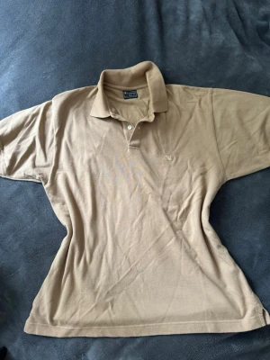 Beige pikétröja från Champion - Klassisk beige pikétröja från Champion med korta ärmar och krage. Tröjan har två knappar vid halsen och diskret broderad Champion-logga på bröstet. Tillverkad i mjuk bomull som känns skön mot huden. Perfekt för en avslappnad och stilren look.