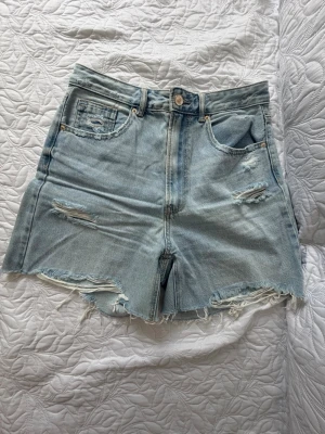 Ljusa denimshorts från Stradivarius - Säljer ett par ljusblå denimshorts från Stradivarius med hög midja och slitna detaljer. Shortsen har råa kanter och klassisk femficksdesign. Perfekta för sommardagar och ger en avslappnad vibe.