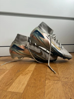 Nike Mercurial Superfly fotbollsskor Elite - Säljer ett par Nike Mercurial Superfly fotbollsskor i ljusgrått med skimrande detaljer i guld och blått. Skorna har hög strumpkrage för extra stöd, snörning och en slitstark ovandel i syntetmaterial. Perfekta för dig som vill ha snabbhet och kontroll på planen.