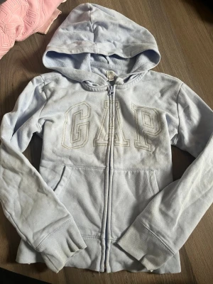Ljusblå hoodie från GAP Kids - Ljusblå hoodie från GAP Kids med dragkedja framtill och stor broderad GAP-logga på bröstet. Hoodien har huva, fickor på sidorna och mjukt fleecefoder på insidan. Perfekt för en chill och avslappnad stil.
