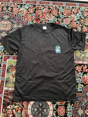Svart t-shirt Casa Santorini Blanca - Svart t-shirt med färgglad tryck på ryggen och mindre tryck på bröstet. Motivet visar vita hus och blå kupoler inspirerade av Santorini, med texten 'Casa Santorini Blanca'. T-shirten har rund hals och korta ärmar, tillverkad i ett mjukt och lätt material.