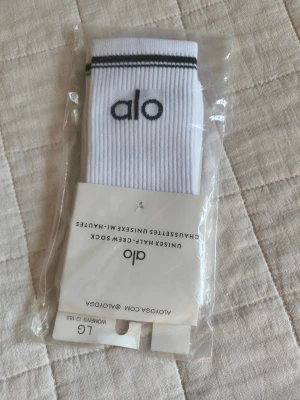 Vita ribbade strumpor från Alo - Säljer ett par vita unisex half-crew strumpor från Alo med svart logga och svarta ränder upptill. Strumporna är ribbade och perfekta för en sportig look. Materialet är mjuk bomullsmix som andas bra.