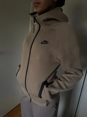 Nike tech hoodie - Beige hoodiejacka från Nike tech med svart logga på bröstet och hel dragkedja framtill. Jackan har hög krage, fickor med svarta detaljer och en sportig look. Perfekt för dig som gillar streetwear och vill ha något bekvämt och stilrent. Aldrig använd