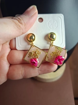 Guldiga örhängen med rosa blomdetalj - Unika örhängen med fyrkantig form i guldton och transparent bakgrund. Dekorerade med en rosa blomma och små guldfärgade detaljer inuti. Perfekta för dig som vill ha något färgglatt och annorlunda i öronen.