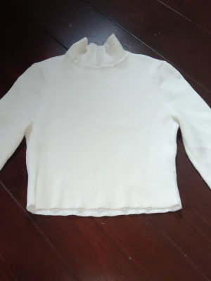 Vit ribbad polotröja - Säljer en enkel och stilren vit stickad polotröja med ribbad struktur. Tröjan har lång ärm och hög krage med lätt vågig kant. Perfekt att matcha med jeans eller kjol för en clean look.