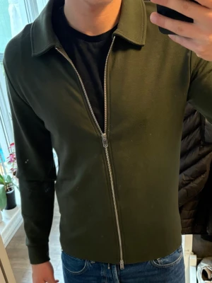 Olivgrön cardiganjacka från Selected - Snygg olivgrön cardiganjacka från Selected med rak passform och hel dragkedja framtill. Jackan har klassisk krage och är tillverkad i ett mjukt, slätt material. Perfekt att slänga över en t-shirt för en clean look.