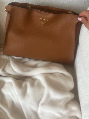 Brun handväska från guess - Snygg brun handväska från guess i slätt, strukturerat läder med guldfärgade metalldetaljer och klassisk trekantslogga framtill. Väskan har en enkel, stilren design och axelrem för smidig användning. Perfekt för dig som gillar lyxiga accessoarer.