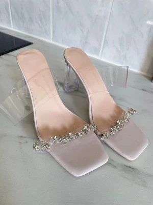 Klara pumps med klack och strass - Säljer ett par trendiga pumps med transparent bred klack och rem. Skorna har fyrkantig tå och dekoreras med gnistrande strass framtill. Färgen är ljusbeige/nude och materialet är syntet med plastdetaljer. Perfekta för dig som vill sticka ut.