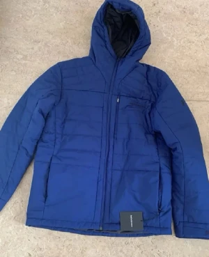 Blå pufferjacka från Peak Performance - Snygg blå pufferjacka från Peak Performance med huva och dragkedja framtill. Jackan har en bröstficka med dragkedja och diskret logga på bröstet. Perfekt för kalla vinterdagar tack vare det vadderade materialet och den sportiga looken.