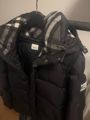 Svart pufferjacka från Burberry - Säljer en svart pufferjacka från Burberry med klassiskt rutigt foder i huvan. Jackan har en vadderad design, dragkedja och tryckknappar framtill samt logotyp på ärmen. Perfekt för kalla dagar. Ärmarna är avtagbara så kan bli en väst också. Fler bilder finns, pris kan diskuteras.