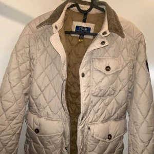 Polo Ralph Lauren jacka - Snygg beige quiltad jacka från Polo Ralph Lauren med klassisk krage i manchester, stora fickor med knappar och dragkedja framtill. Jackan har ett rutigt mönster och är perfekt för dig som vill ha en stilren och tidlös look nu till våren. Den är också perfekt för grishstilen. Tags tillkommer. Den är i storlek S och är i nyskick. Nypris 5195kr säljs för 1999kr. Den är slutsåld överallt, passa på o plicka hem den nu!