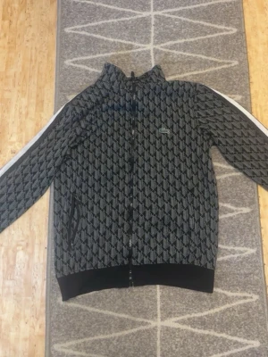 Mönstrad bomberjacka från Lacoste - Snygg bomberjacka från Lacoste med svart och grått geometriskt mönster, vita ränder längs ärmarna och dragkedja framtill. Jackan har ribbade muddar och hög krage, perfekt för en sportig och trendig look.