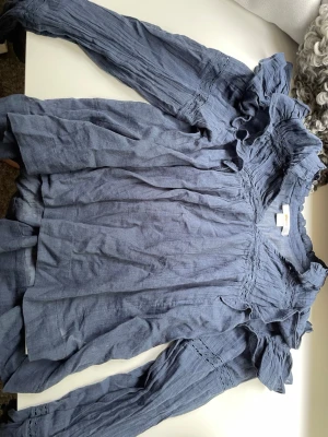 Blå volangblus från Gina Tricot - Supersöt blå blus från Gina Tricot med volangdetaljer vid axlar och hals. Blusen har långärmade ärmar och är tillverkad i ett lätt, vävt tyg med fina broderade detaljer. Perfekt för dig som gillar en romantisk och trendig stil.