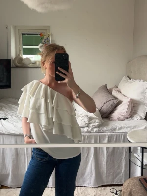 Vit oneshoulder topp med volanger - Säljer en superfin vit oneshoulder topp med flera lager volanger. Toppen har en asymmetrisk design och är perfekt för dig som vill sticka ut lite extra. Materialet känns mjukt och lätt, och toppen är ärmlös på ena sidan. Passar perfekt till jeans eller kjol.