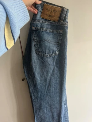 Blå raka jeans från Rocky - Säljer ett par klassiska blå jeans från Rocky med rak passform och hög midja. Jeansen har fem fickor, knappgylf och tydliga sömmar. Perfekta för en avslappnad och tidlös stil.