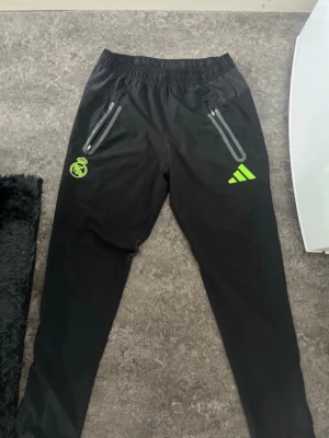 Svarta Real Madrid mjukisbyxor Adidas slim fit - Svarta mjukisbyxor från Adidas med Real Madrid-logga och neongröna detaljer. Byxorna har dragkedjor vid fickorna och smal passform med resår i midjan. Perfekta för chill eller träning, med sportig look och bekvämt material.