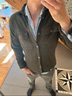 Mörkgrön overshirt från Jack & Jones - Stilren mörkgrön overshirt från Jack & Jones i mjukt tyg. Två bröstfickor med lock och knappar framtill. Perfekt att bära över en skjorta för en avslappnad men snygg look. Klassisk krage och raka ärmar. Perfekt nu inför sommaren.