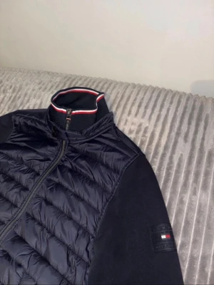 Svart pufferjacka från Tommy Hilfiger - Snygg svart pufferjacka från Tommy Hilfiger med quiltad framsida och dragkedja. Jackan har ribbade muddar, hög krage med röd, vit och blå detalj samt logga på ärmen. Perfekt för kyliga dagar och enkel att matcha.