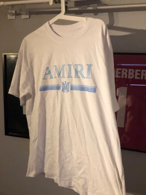Vit AMIRI t-shirt med blått tryck - Stor i storlek och sitter typ som en Large! Säljer en vit t-shirt från AMIRI med stort blått tryck på bröstet och klassisk rund hals. T-shirten har korta ärmar och är tillverkad i mjuk bomull för en skön och avslappnad look. Perfekt för dig som gillar streetwear och stilrena plagg.