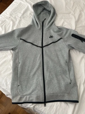 Grå Nike Tech Fleece hoodie - Snygg grå Nike Tech Fleece hoodie med hel dragkedja och svarta detaljer. Har huva, två sidofickor och en extra ficka med dragkedja på ärmen. Tillverkad i mjukt och lätt material som passar perfekt till streetwear-stilen. Nike-logga på bröstet.