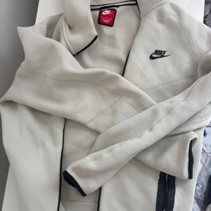 Beige Nike zip-hoodie i bomull - Snygg beige nike tech hoodie från Nike med svart logga på bröstet och svarta detaljer längs dragkedjan. Jackan har huva och är tillverkad i mjuk bomull, perfekt för chill dagar eller när du vill ha en sportig look. Priset kan diskuteras.