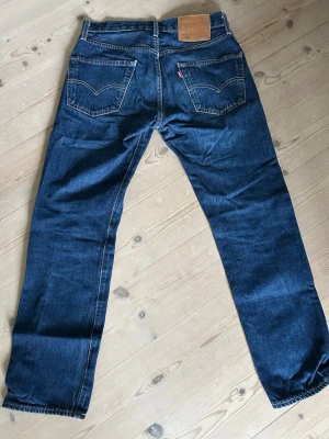 Levi's jeans - Säljer ett par klassiska blå Levi's jeans med raka ben. Jeansen har en mörk tvätt, kontrastsömmar och den ikoniska läderlappen bak i midjan. 