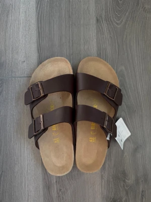 Bruna Birkenstock sandaler med spännen - Säljer ett par klassiska Birkenstock sandaler i mörkbrunt läder med två breda remmar och justerbara metallspännen. Fotbädden är i mjukt mockamaterial och sulan är ergonomiskt utformad för extra komfort. Perfekta för en avslappnad och stilren look.