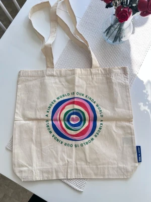 Tygväska med färgglatt tryck (ny) - En större beige tygväska med ett färgglatt tryck på framsidan. Köpt på Cotton On i Australien. Oanvänd.  Bandens längd: 77-78cm.  Storlek: 45x42cm
