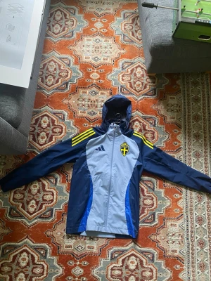 Blå Adidas vindjacka Sverige - Snygg blå regnjacka från Adidas med Sveriges landslagsemblem på bröstet och gula detaljer på axlarna. Jackan har huva, dragkedja framtill och meshfoder på insidan. Perfekt för dig som gillar sportig stil och vill visa ditt stöd för Sverige. Använt några fåtal gånger.
