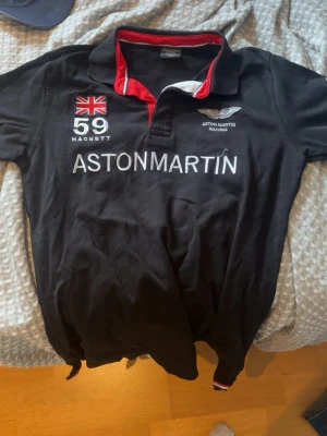Svart Aston Martin Racing piké - Snygg svart pikétröja från Hackett x Aston Martin Racing med broderad brittisk flagga, nummer 59 och tydlig Aston Martin-logga på bröstet. Kragen har röda och vita detaljer som ger en sportig look. Perfekt för dig som gillar racing och stil. M/L i storlek, Pris kan diskuteras:)