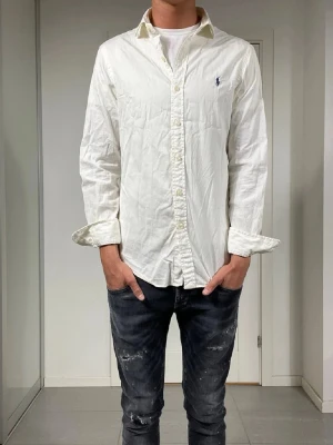 Vit skjorta från Ralph Lauren - Klassisk vit skjorta från Ralph Lauren med broderad logga på bröstet. Skjortan har lång ärm, knappar framtill och en avslappnad passform. Perfekt till jeans eller chinos för en clean och stilren look.
