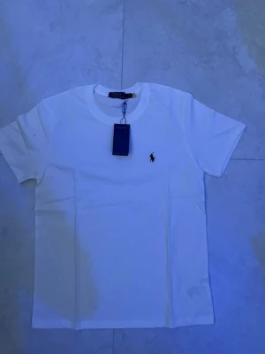 Vit t-shirt från Polo Ralph Lauren - Klassisk vit t-shirt från Polo Ralph Lauren med rund hals och korta ärmar. Liten broderad logga i svart på bröstet. Tillverkad i mjuk bomull för en bekväm känsla och perfekt passform.