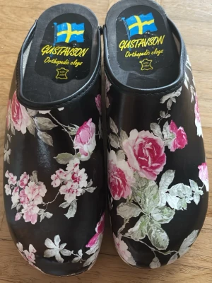 Blommiga svarta trätofflor Gustavson - Säljer ett par svarta trätofflor från Gustavson (ortopedin clogs). Tofflorna har ett snyggt blommönster i svart, rosa, vitt och grönt som ger en somrig och unik look. 