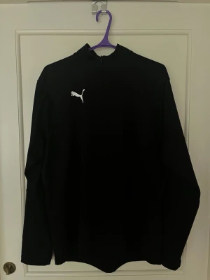 Svart träningströja 1/4 zip från Puma strl L - Svart långärmad träningströja från Puma med halv dragkedja och vit Puma-logga på bröstet. Tillverkad i ett stretchigt funktionsmaterial som passar perfekt för träning eller uppvärmning. Storlek L.