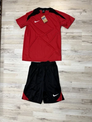 Nike röd och svart träningsset - Säljer ett sportigt träningsset från Nike med röd t-shirt och svarta shorts. T-shirten har svarta detaljer vid axlarna och ett vitt Nike-märke på bröstet. Shortsen är svarta med röda detaljer och har Nike-logga på benet. Perfekt för fotboll eller gym.