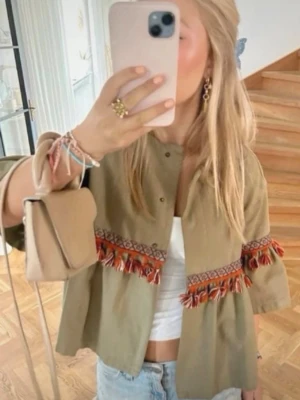  overshirt med fransar och mönster - Säljer en  overshirt med unika röda och orange fransdetaljer samt broderat mönster runt ärmar och midja. Jackan har en loose passform, knappar framtill och trekvartsärmar. Perfekt för dig som vill sticka ut med en bohemisk touch.