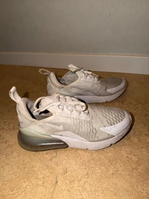 Nike Air Max 270 vita sneakers - Nike Air Max 270 sneakers i vitt med mesh-överdel och synlig Air-enhet i hälen. Skorna har en sportig design med snörning, dragflik bak och diskreta loggor på sidan och plösen. Perfekta för dig som gillar streetwear och bekväma skor.