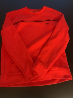 Röd sweatshirt från BOSS - Riktigt snygg röd sweatshirt från BOSS med svart logga på bröstet och en tunn svart rand över bröstet. Tröjan har rund halsringning och långa ärmar. Perfekt för dig som vill ha en enkel men stilren look.