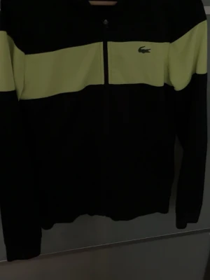 Svart/limegrön hoodie Lacoste  - Snygg hoodie från Lacoste i svart med bred limegrön panel över bröstet och armarna. Dragkedja framtill och klassisk Lacoste-krokodil på vänster sida. Tillverkad i mjukt och bekvämt material, perfekt för en sportig och avslappnad stil. Pris kan diskuteras.