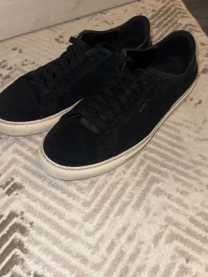 Svarta sneakers från Axel Arigato - Stilrena svarta sneakers från Axel Arigato i mocka med svart läderdetalj vid hälen och vit sula. Klassisk låg modell med svarta skosnören och diskret logga i guld på sidan. Perfekta för en clean och modern look.