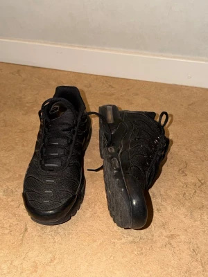 Svarta Nike Air Max Plus sneakers - Säljer ett par helsvarta Nike Air Max Plus sneakers med klassisk mesh- och syntetöverdel, synlig Air-dämpning i sulan och vågiga detaljer på sidorna. Skorna har en sportig look och är perfekta för dig som gillar streetwear.