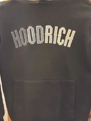 Svart hoodie från Hoodrich - Säljer en svart hoodie från Hoodrich med stor logga i grått och vitt på bröstet. Klassisk känguruficka framtill och huva. Perfekt för dig som gillar streetwear och vill ha en stilren men ändå statement tröja. Använd skick XS