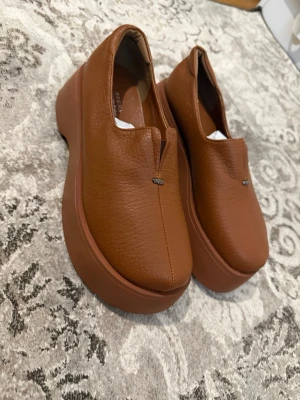 Bruna platåloafers i skinn - Säljer ett par bruna loafers med chunky platåsula och stilren design. Skorna är tillverkade i slätt skinn och har en liten metall-detalj framtill. Perfekta för dig som vill ha en modern och trendig look.